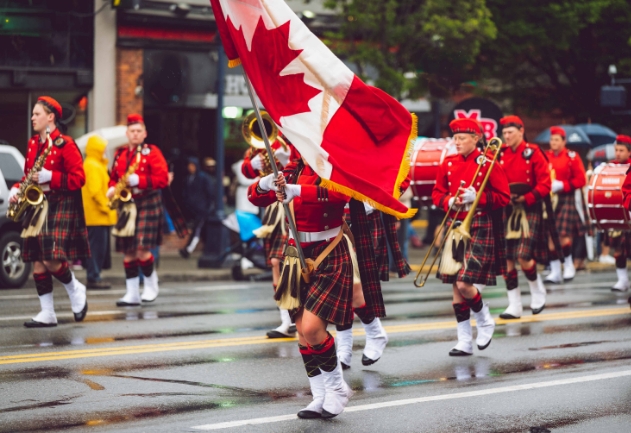 Membros de uma fanfarra, segurando a bandeira do Canadá atravessendo uma rua tocando os seus diversos instrumentos de sopro e percussão