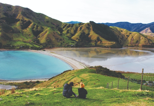 Duas pessoas sentadas na grama admirando uma paisagem composta por um lago com montanhas ao seu redor