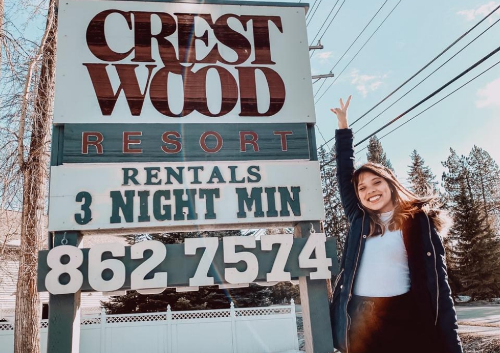 Julia França ao lado de uma placa do 'Crestwood Resort', à luz do dia, com árvores ao fundo