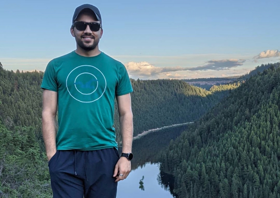 Heitor Neiva parado de costas a uma paisagem composta por um rio que se adentra por entre uma densa floresta, sob um céu limpo, com poucas e distantes nuvens ao fundo