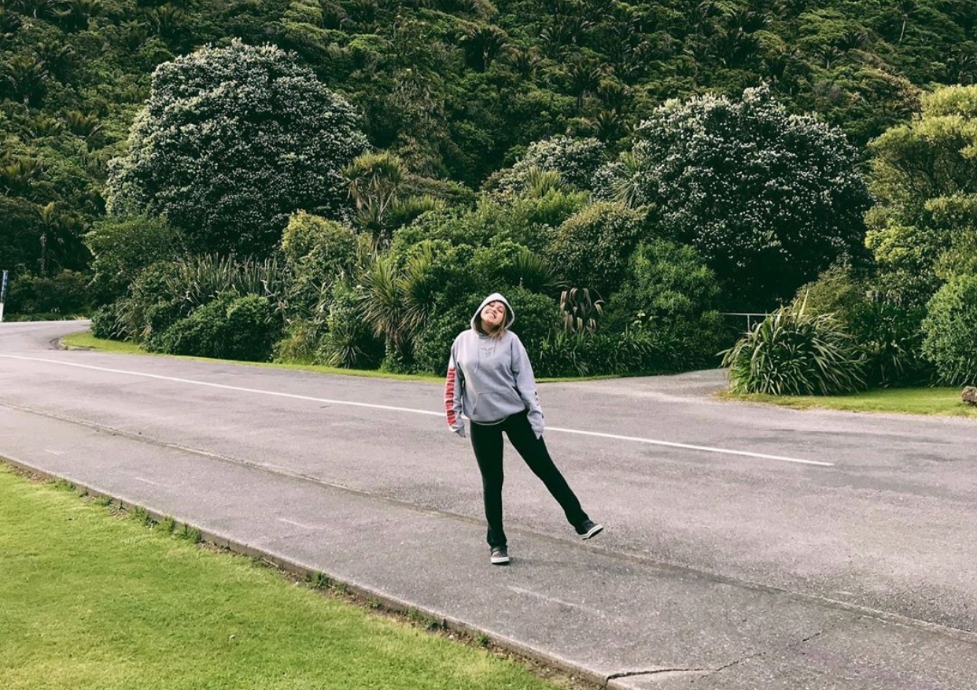 Lia Gomes sorrindo, parada em uma rua vazia, com árvores e vegetação rasteira ao fundo, sob à luz do dia